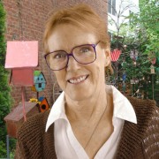 Woman with short red hair and glasses standing in front of a red brick wall with birdhouses and other decorations nearby